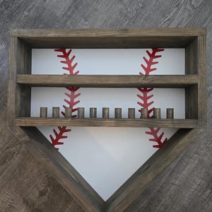 Puede incluir: Estante de madera con temática de béisbol en forma de home plate. El estante tiene un fondo blanco con detalles de costuras de béisbol rojas. Cuenta con dos estantes y pequeñas clavijas de madera para exhibición.