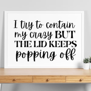 May include: A framed print with the text "I try to contain my crazy BUT THE LID KEEPS popping off" in black script. The print is in a white frame, set on a wooden desk with a small potted plant.