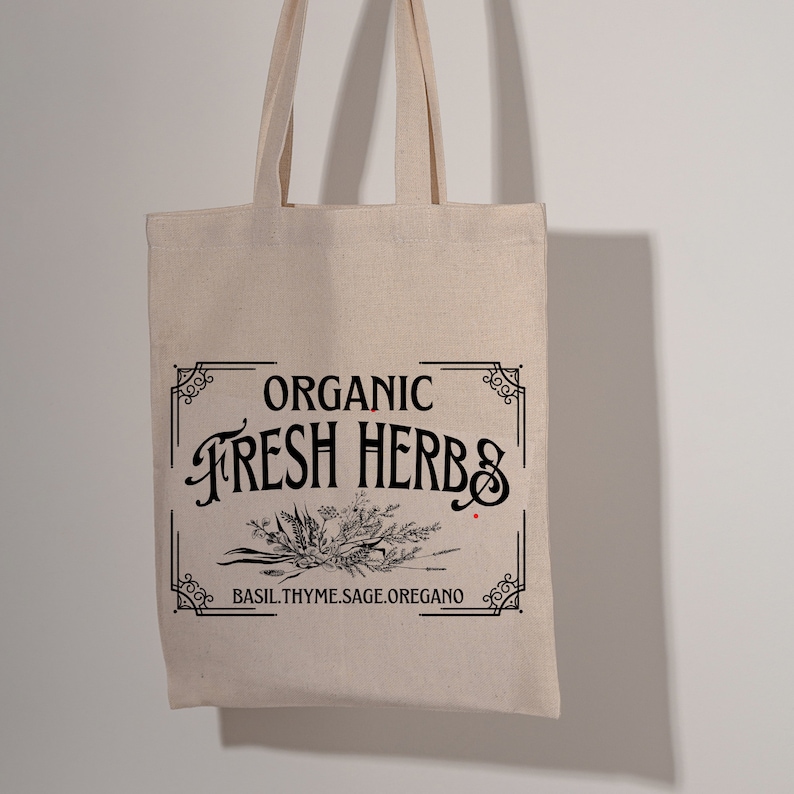 May include: A beige canvas tote bag with black text and a decorative border. The text reads "ORGANIC FRESH HERBS" above a botanical illustration and the words "BASIL.THYME.SAGE.OREGANO" below. The bag has two handles.