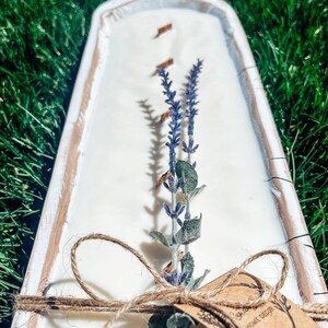 May include: A white rectangular candle in a wooden tray, decorated with dried lavender and a twine bow. The candle has three wooden wicks and a Home Decor tag. The tray is approximately 12 inches long.