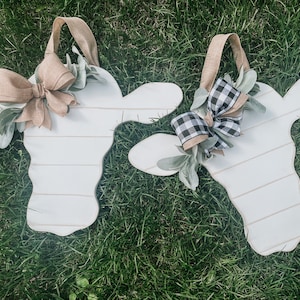 May include: Two white cow-shaped wooden wall hangings with horizontal lines. One features a solid tan burlap bow, the other a black and white buffalo check bow. Both are adorned with green faux leaves and include a tan burlap hanging strap.