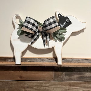 May include: A white wooden pig cutout featuring a black and white buffalo check ribbon bow, green faux leaves, and a black ear tag with the white text "Seeck" written in a cursive script. The pig has a distressed finish along the edges.