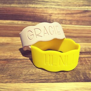 May include: Two silicone bracelets with wavy edges. One bracelet is light pink with the name GRACIE embossed in capital letters. The second bracelet is bright yellow with the name HENLE embossed in capital letters on the front surface.