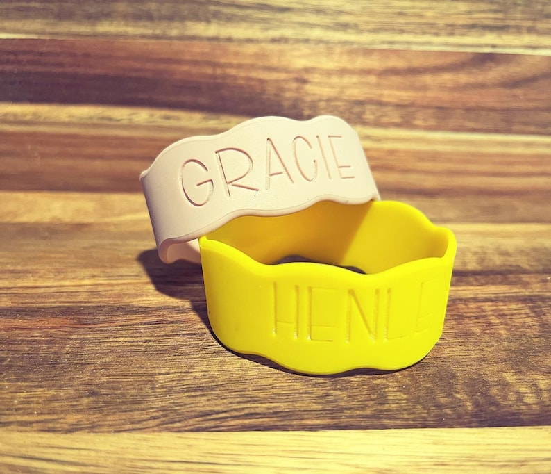May include: Two silicone bracelets with wavy edges. One bracelet is light pink with the name GRACIE embossed in capital letters. The second bracelet is bright yellow with the name HENLE embossed in capital letters on the front surface.