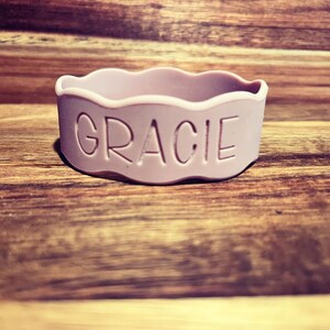 May include: A light lavender silicone bracelet featuring a wavy, scalloped top edge and the name GRACIE debossed in capital letters on the front surface. The band is circular and has a smooth, matte finish throughout its entire exterior structure.