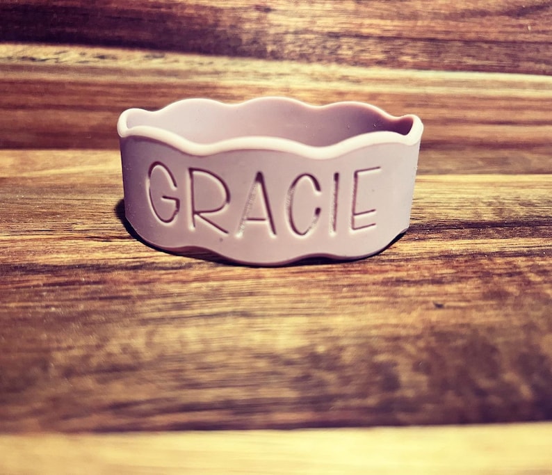 May include: A light lavender silicone bracelet featuring a wavy, scalloped top edge and the name GRACIE debossed in capital letters on the front surface. The band is circular and has a smooth, matte finish throughout its entire exterior structure.