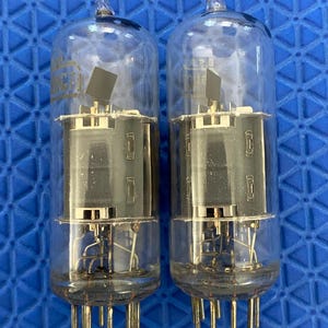May include: Two clear glass vacuum tubes featuring internal metal components and a series of metallic pins at the base. Each cylindrical tube contains a dark rectangular plate structure and delicate wire connections visible through the transparent glass.