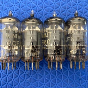 May include: Four vintage vacuum tubes with glass enclosures and metal pins. Each tube has internal components visible through the clear glass. Text on the tubes includes "PHILIPS", "PCF80", and "F80". The tubes are on a blue surface.