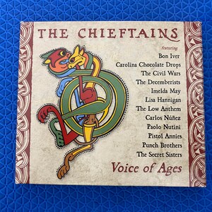 Può includere: Custodia di un CD per l'album "Voice of Ages" di "The Chieftains". La copertina presenta un colorato disegno a nodo celtico con testo che elenca artisti come Bon Iver e The Civil Wars. Lo sfondo è beige chiaro con un bordo marrone.