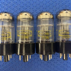 May include: Four vintage vacuum tubes with clear glass bulbs and black bases. Each tube has the "RAYTHEON REGISTERED" logo in yellow. The tubes are arranged in a row against a blue background, showcasing their design and branding.