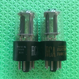 May include: Two vintage RCA vacuum tubes with clear glass enclosures and black bases. The bases have the RCA logo and text that reads "RADIOTRON ELECTRON TUBE" and "MADE IN U.S.A."