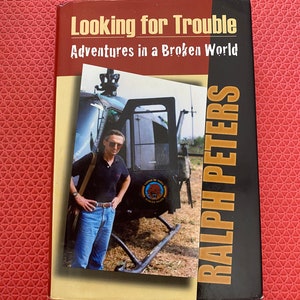 May include: A hardcover book titled "Looking for Trouble: Adventures in a Broken World" by Ralph Peters. The cover features a photo of a man in front of a helicopter. The book has a red and tan color scheme.