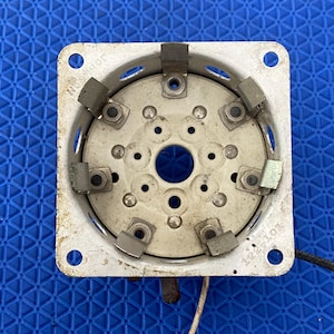 May include: A square, off-white metal vacuum tube socket component featuring a circular center with multiple pin holes, surrounded by six metallic contact clips. The frame is stamped with the text Johnson and the numerical sequence 122-101.