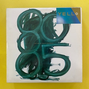 May include: A square album cover with a white background and large, abstract green brushstrokes. The upper right corner features a sticker with the word "YELLO" in gold lettering. The album title is "YELLO 3000 THE NEW MIX IN ONE GO".