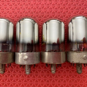 May include: Four vintage vacuum tubes with clear glass bulbs and silver-colored metal bases. The tubes have a cylindrical shape and are arranged in a row. The bases have pins for electrical connections. The background is a red textured surface.