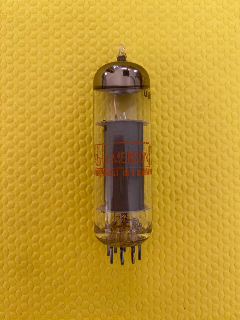 Puede incluir: Tubo de vac&iacute;o vintage con una carcasa de vidrio transparente y una parte superior plateada. El tubo tiene componentes internos y una estructura cil&iacute;ndrica de color gris oscuro. El texto "EMERSON" es visible. El tubo tiene m&uacute;ltiples pines en la base.