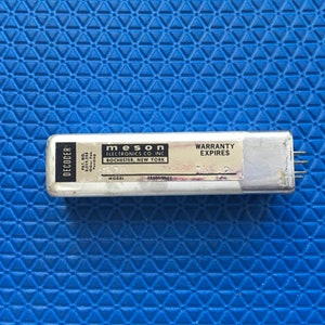 May include: A silver-colored electronic decoder with the text "DECODER" on the left side. The label reads "MESON ELECTRONICS CO. INC. ROCHESTER, NEW YORK" and "WARRANTY EXPIRES". The device has several pins on the right side. The background is a blue, textured surface.