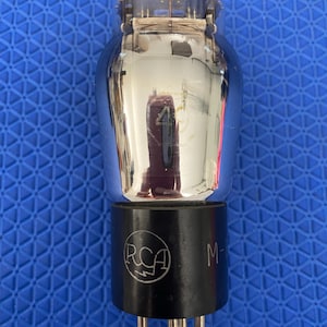 May include: A vintage vacuum tube with a clear glass bulb and a shiny metal interior. The black base features the RCA logo and the letter "M-". The tube has several pins at the bottom.