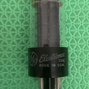 May include: A clear glass vacuum tube with a black base featuring the GE logo and the text Electronic TUBE MADE IN U.S.A. The internal components are dark grey and metallic, with multiple metal pins extending from the bottom of the black base.