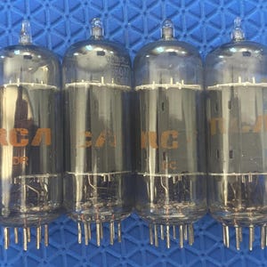 May include: Four vintage RCA vacuum tubes with clear glass enclosures and silver pins. The tubes have the RCA logo in orange lettering. The background is a textured blue surface.