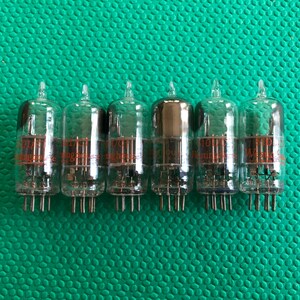 Puede incluir: Seis válvulas de vacío vintage con carcasas de vidrio y múltiples pines en la base. Los tubos tienen componentes internos y texto naranja. Están dispuestos en fila sobre una superficie verde texturizada.