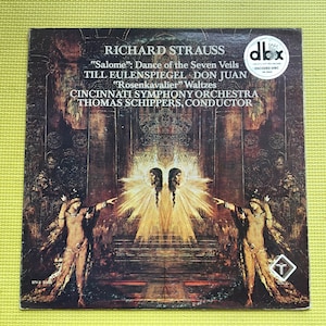 Puede incluir: Una portada de álbum de obras de Richard Strauss, incluyendo "Salomé" y "Don Juan", interpretadas por la Orquesta Sinfónica de Cincinnati. La obra de arte presenta una ilustración oscura y ornamentada con figuras y texto. El álbum es un disco codificado dbx.