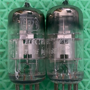 May include: Two vintage vacuum tubes with clear glass enclosures. Each tube has a silver metal interior with the number "5879" visible. The tubes have multiple pins at the base and a pointed glass tip at the top. The background is a green surface.