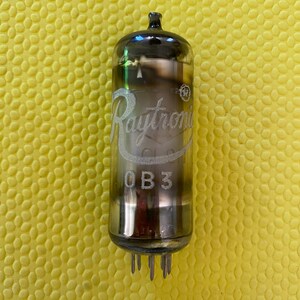May include: A vintage Raytronic OB3 vacuum tube with a clear glass body and silver-colored top. The tube has a series of pins at the base and the Raytronic logo is visible. The tube is set against a yellow textured background.