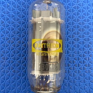 May include: A vintage Raytheon 60KD6 valve. The glass tube has a metal base with several pins. The Raytheon logo is in a yellow oval, with the word "REGISTERED" below it. The valve is clear with internal components visible.