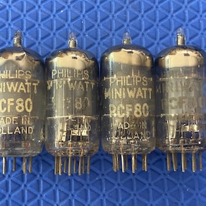 May include: Four vintage Philips Miniwatt PCF80 vacuum tubes. Each tube is clear glass with a silver top and multiple pins at the base. Text on the tubes reads "PHILIPS MINI WATT PCF80 MADE IN HOLLAND".