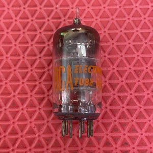 Peut inclure: Un tube à vide RCA en verre transparent avec le texte orange "RCA ELECTRON TUBE". Le tube a un sommet pointu et plusieurs broches à la base. Le fond est une surface rouge texturée.