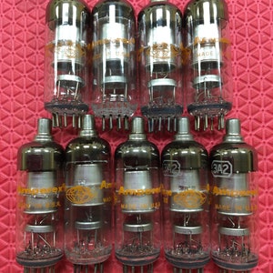 Puede incluir: Diez tubos de vacío de vidrio transparente con tapas plateadas y bases marrones están dispuestos sobre una superficie roja. Los tubos tienen componentes internos y el texto "Amperex" y "Made in U.S.A." es visible.