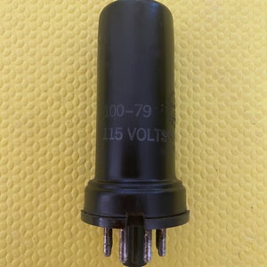 Puede incluir: Un componente electrónico negro con el texto "100-79" y "115 VOLTS". Tiene forma cilíndrica y pines metálicos en la base. El fondo es una superficie amarilla texturizada.