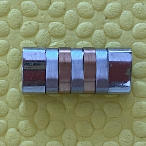 May include: A rectangular metal object with alternating silver and copper-coloured segments. The object is composed of six segments, with the end segments being silver. The object is resting on a textured yellow surface.