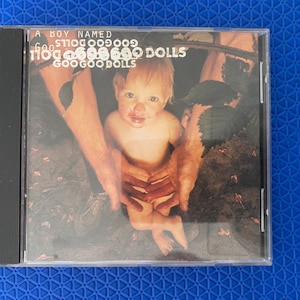 May include: CD case for the Goo Goo Dolls album, "A Boy Named Goo." The cover art features a baby held by two sets of hands. The album title and band name are printed in white text.