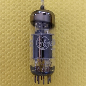 May include: A vintage vacuum tube with a glass enclosure and metal components. The tube has a cylindrical shape with a rounded top and a base with multiple pins. The GE logo is visible on the internal components. The tube is designed for electronic devices.