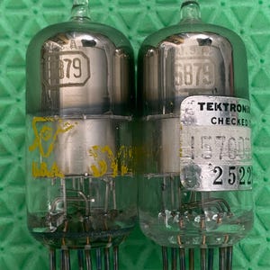Puede incluir: Dos válvulas de vacío vintage con carcasas de vidrio y bases metálicas. Cada válvula tiene una etiqueta con el número "6879" y una pegatina "Tektronix Checked". Las válvulas están sobre una superficie verde.