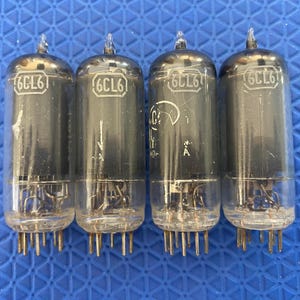 May include: Four vintage 6CL6 vacuum tubes. Each tube has a glass enclosure with a metal top and a base with pins. The tubes are arranged in a row against a blue background.