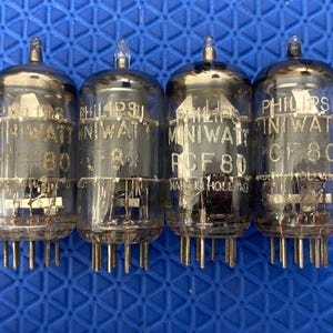May include: Four vintage Philips Miniwatt RCF 80 vacuum tubes. Each tube is clear glass with a metal base and multiple pins. The tubes have the Philips Miniwatt logo and "Made in Holland" printed on them. The tubes are on a blue surface.