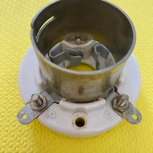 May include: A vintage light fixture component with a cylindrical metal housing and a white ceramic base. The base has two metal terminals with screws and the letters "P" and "F" stamped on it. The fixture is set against a yellow background.