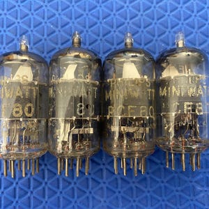 May include: Four vintage Philips Miniwatt vacuum tubes. Each tube is clear glass with a silver top and a base with multiple pins. The tubes have printed text including "80" and "CE80". The background is a blue surface.
