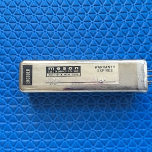 May include: A rectangular metal encoder with the text "ENCODER" on the left side. The label reads "meson ELECTRONICS CO INC ROCHESTER, NEW YORK" and "WARRANTY EXPIRES". The model number is J0101 and the frequency is 77.0.