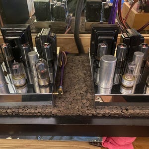 May include: Close-up of two vintage audio amplifiers with multiple vacuum tubes and transformers. The amplifiers are mounted on a dark surface with a textured, cork-like material. The components are silver and black.