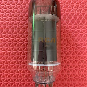 May include: A vintage vacuum tube with a clear glass enclosure. The tube has internal components and metal pins at the base. The text "RCA" and "DH" are printed on the internal components. The tube is cylindrical in shape.