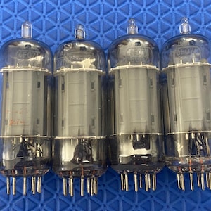 May include: Four vintage vacuum tubes with clear glass bulbs and metal bases. Each tube has a cylindrical shape with internal components visible. The tubes are labeled "U.S.A. 6F07" and are arranged against a blue textured background.