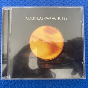 Könnte beinhalten: Eine standardmäßige CD-Hülle aus durchsichtigem Kunststoff mit dem Album Parachutes von Coldplay. Das Cover zeigt einen schwarzen Hintergrund mit einem großen, leuchtend gelben Globus in der Mitte und dem Text COLDPLAY PARACHUTES in Weiß.