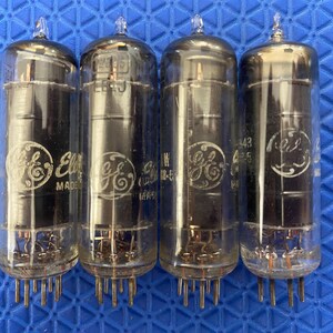 May include: Four vintage vacuum tubes with clear glass bodies and metal pins. Each tube has a rounded top and a cylindrical shape, with visible internal components. The tubes are marked with the "GE" logo and other text, likely model numbers.