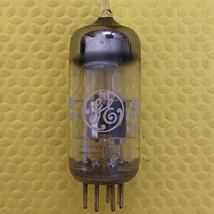 May include: A vintage vacuum tube with a clear glass enclosure and a GE logo. The tube has a cylindrical shape with a pointed top and multiple pins at the base. The background is a textured yellow surface.