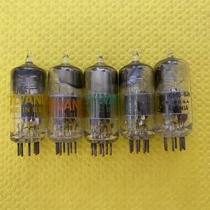May include: Five vintage vacuum tubes with clear glass bodies and black internal components are arranged in a row. Each tube has a label with the brand name "SYLVANIA" in various colors. The tubes are on a yellow surface.