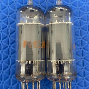 May include: Two vintage vacuum tubes with clear glass enclosures. Each tube has a cylindrical shape with internal components visible. The tubes have metal pins at the base and a small glass tip at the top. The text "U.S.A. OF 07" is visible on the glass.